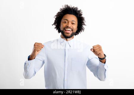 Excited young african american guy in formal white shirt posing isolated on white background, raising fists up in winner gesture, copy space Stock Photo