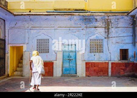 An old man walking with a wooden stick on a public place with buildings in various colours Stock Photo