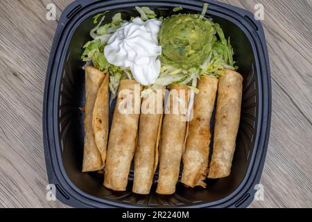 Overhead view of side order of deep fried, crispy taquitos packed in a to go box with large scoops of sour cream and guacamole. Stock Photo