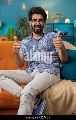 Like. Happy excited indian man looking approvingly at camera showing thumbs up, like sign positive something, good great news, positive feedback. Young hindu guy sitting on couch at home living room Stock Photo