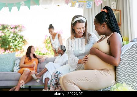 This feels so wonderful. a young woman feeling her friends baby move in her belly during her baby shower. Stock Photo