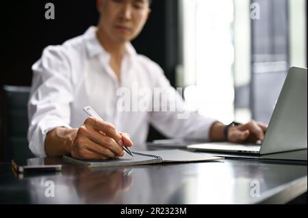 Close-up image of a professional millennial Asian businessman or male boss in a white shirt working at his desk, writing or taking notes on his notebo Stock Photo