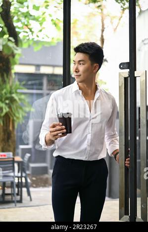 Portrait of a handsome and happy millennial Asian businessman with his coffee cup opening the door, entering or leaving a coffee shop. Stock Photo