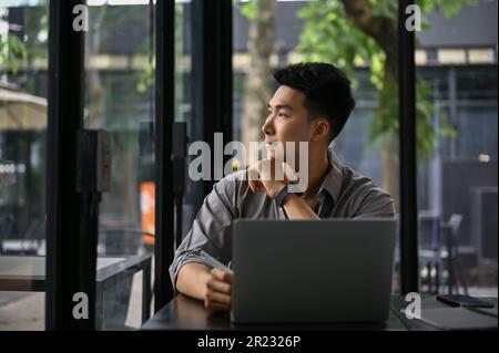 Inspired and handsome millennial Asian businessman thinking and daydreaming while looking out the window in the cafe. Stock Photo