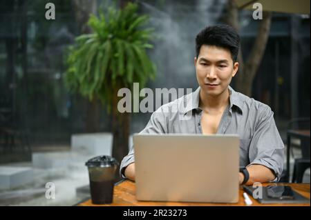 Handsome millennial Asian man is using his laptop computer, working on his work, and sitting at a coffee shop in an outdoor space. Stock Photo