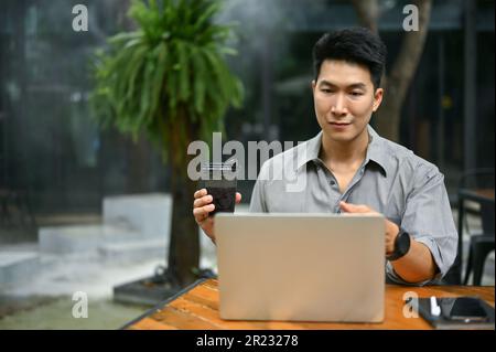 Handsome and smart millennial Asian businessman having an online meeting with his team while remote working at a coffee shop. Stock Photo