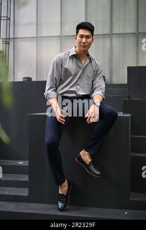 Portrait of a handsome millennial Asian businessman sits outside with his takeaway coffee cup. Stock Photo