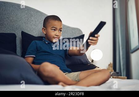 Tv can be a helpful tool in learning. a young boy using a remote control while lying on his bed. Stock Photo