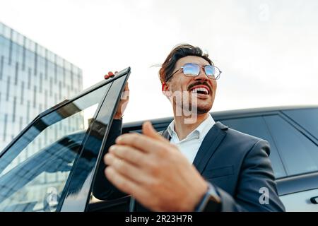 Furious driver gesticulates with open hand on white car. Desperate young  man with sunglasses driving with anger. Upset, aggressive, impatient, bad  manners concepts. Cold blue effect on sunny day Stock Photo