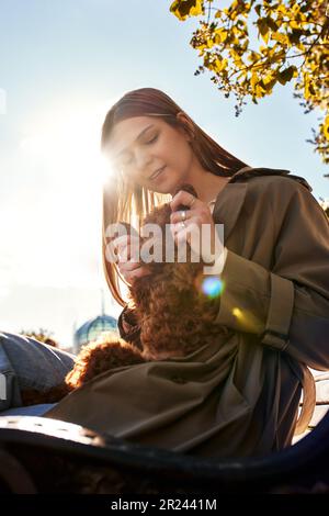 A young smiling blonde girl in a stylish beige raincoat holds and hugs her pet toy poodle in her arms. The girl and the dog are resting in the park. S Stock Photo