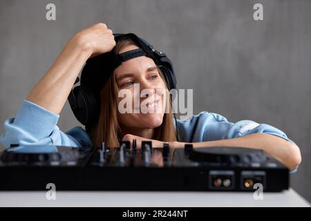 Close-up portrait of a young pretty long-haired DJ girl in a blue sweater and black baseball cap mixing tracks using a DJ console. Music, fun and crea Stock Photo