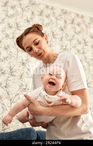 Loving mom carying her little baby at home. Bright portrait of happy mom holding her infant child on hands. Mother hugging her little baby Stock Photo