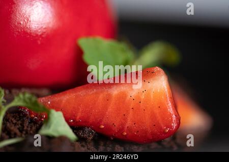 Selective focus of a strawberry on the brownie crumbs. Sphere curd cake with smooth surface and mirror glaze. Red dessert. Stock Photo