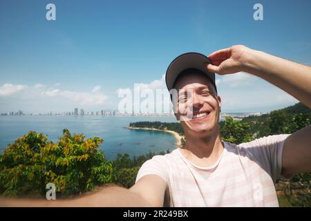 Happy man taking selfie photo from summer vacation day. Handsome tourist wearing cap and smiling at camera against landscape with beach. Stock Photo