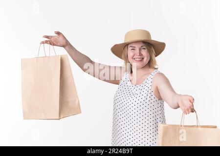 Woman shopper in white dress with black polka dots and straw hat holds craft paper shopping bags in hands on white background Stock Photo