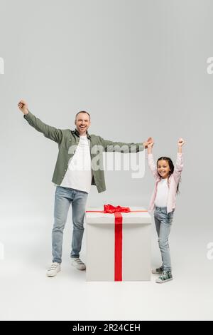 Full length of excited and carefree father and preteen daughter looking at camera near big present during child protection day celebration on grey bac Stock Photo