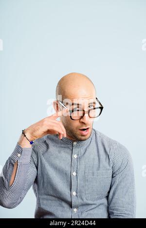 Oh, I see you alright. a handsome young man sitting alone in the studio and pulling funny faces while wearing glasses. Stock Photo