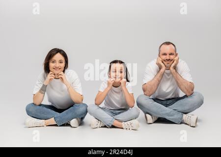 joyous father and tattooed mother with short hair and cheerful preteen daughter sitting with crossed legs in white t-shirts and blue denim jeans on gr Stock Photo