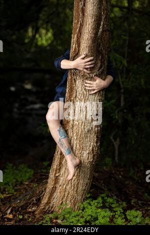 A young female stands in a forest, her arms wrapped around a tree trunk in a tender embrace Stock Photo