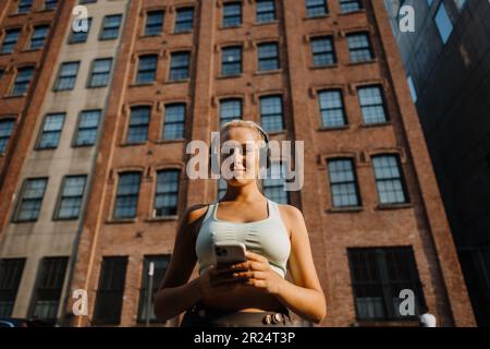 Sportive young blonde woman in fitness clothing wearing headphones and turning music on smartphone Stock Photo