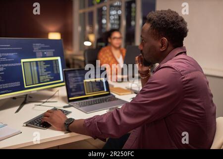 African American developer writing computer codes on computer while working at his workplace in office till late night Stock Photo