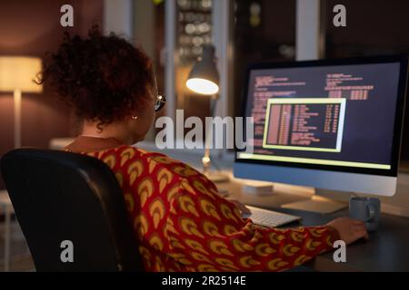 Rear view of female programmer sitting at her workplace in front of computer monitor and working with codes till late night Stock Photo