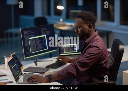African American developer working with computer program on computer while sitting at his workplace in late evening Stock Photo