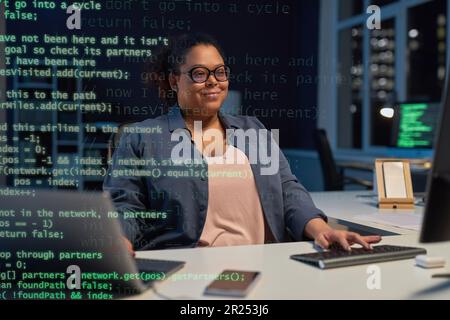 African American woman typing codes on computer while sitting at her workplace in dark IT office Stock Photo