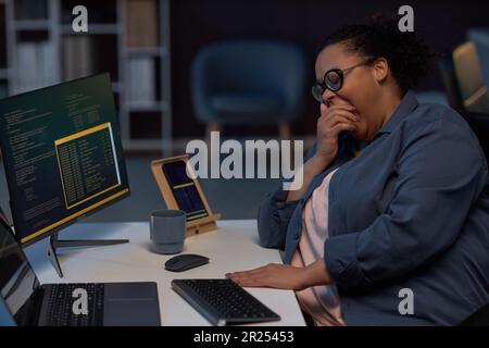Tired programmer yawning while working at her workplace on computer till late night during deadline Stock Photo