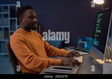 African American developer working with security codes of program on computer sitting at his workplace in dark IT office Stock Photo
