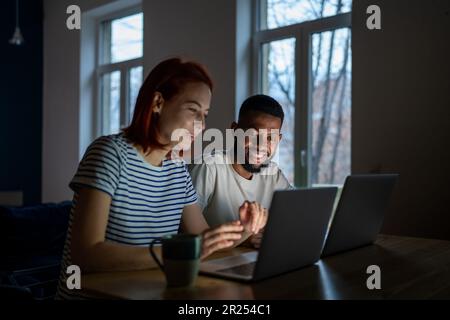 Smiling multiracial family couple enjoy freelancing, sitting at table working on laptops together Stock Photo