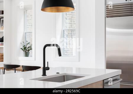 A kitchen sink detail with a black faucet under a black and gold pendant light, white oak island, white countertop, and decorations in the background. Stock Photo