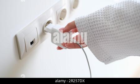 Woman in white bathrobe inserting plug into outlet at home with hand closeup Stock Photo
