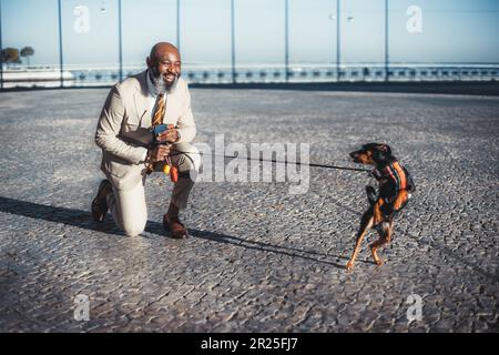 Selective focus on a bald bearded man with a dog, has one knee on Lisbon's cobblestone pavement and is smiling at the dog that is behaving funny; Hold Stock Photo