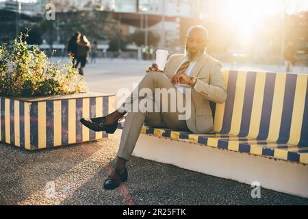 Lisbon; A well-dressed bald unshaven mature black man sitting on a wooden bench with yellow and blue vertical stripes; man checks the time on his wris Stock Photo