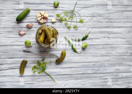Pickled cucumbers in an open jar, gherkin on a fork and garlic on a light wooden background, top view, copy space. Homemade canned cucumbers. Stock Photo