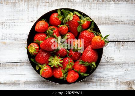 Large ripe juicy berries of organic strawberries in a black round plate on a light wooden background, top view. Stock Photo