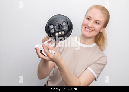 Smiling Caucasian Woman Holds IP Smart Outdoor Security Camera On White Grey Background. Smart Home, House Surveillance, System With Optical Zoom Stock Photo