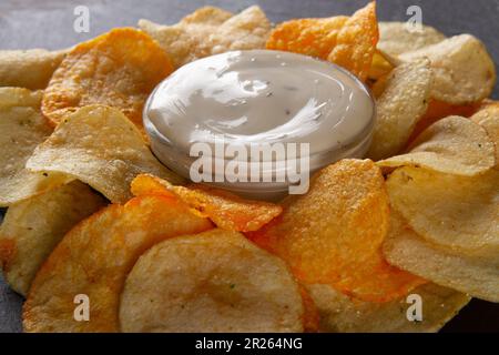 Close-up Side view of heap of potato chips with creamy white dipping sauce on black slate serving board on grey plaster table. Chic Snacking Board Stock Photo