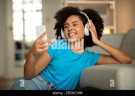 Glad young black curly woman in wireless headphones looks at phone, enjoy weekend, listen music Stock Photo