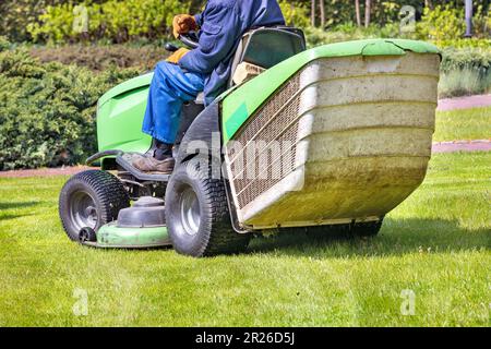 A worker in blue overalls drives a tractor lawn mower and mows the green grass of the lawn. on a blurred background of a spring garden. Copy space. Stock Photo