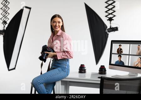 Happy creative photographer lady sitting on table workplace, holding her DSLR camera, working in modern photostudio Stock Photo