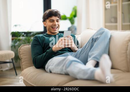 Happy Middle Eastern Young Man Using Cellphone Laughing At Home Stock Photo