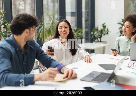 Three Diverse Coworkers Chatting Sitting At Table In Modern Office Stock Photo