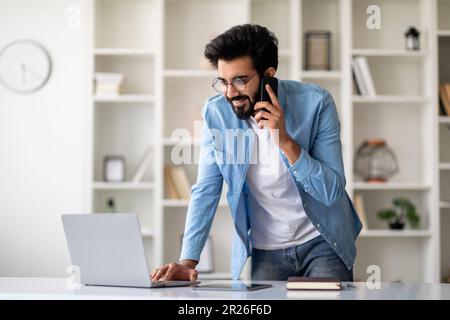 Young Indian Man With Cellphone And Laptop Working Remotely From Home Office Stock Photo