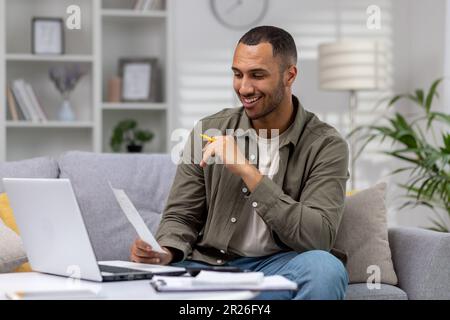 Young successful hispanic man at home sitting on sofa in living room, calculating and paying bills, man with calculator and laptop online paying for r Stock Photo