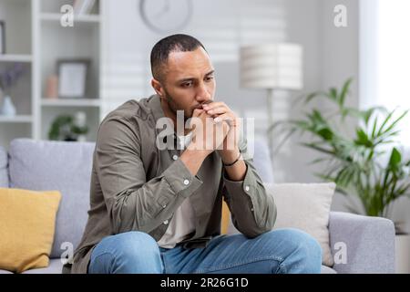 Upset man sitting alone at home, depressed hispanic man on sofa in living room in casual clothes thinking about difficult decision. Stock Photo