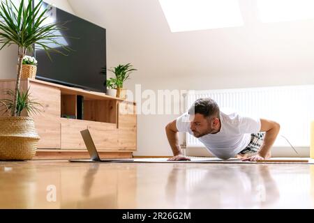 Man doing push-ups on the floor at home, doing plank, watching online workout training in the internet. Stock Photo