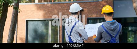 Making the best house. Rear view of two builders standing outdoors with an open blueprint discussing the half completed cottage house Stock Photo