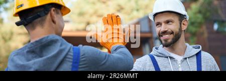 Leave It to the Professionals. Two positive young male engineers in hard hats smiling at each other, giving high five while working on cottage Stock Photo